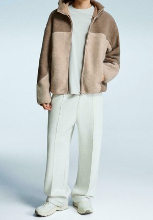 Personne portant une veste polaire beige et marron à fermeture éclair, un pantalon large crème, des baskets blanches, debout avec une main dans la poche.