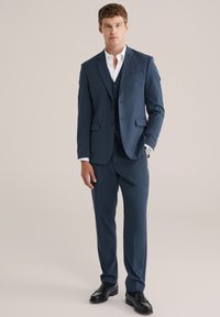Mann in einem marineblauen Dreiteiler mit weißem Hemd und schwarzen Slippern vor einem schlichten beige Hintergrund.