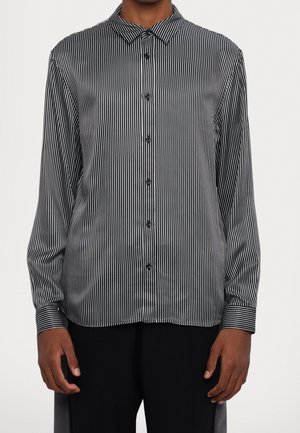 Man draagt een zwart-wit gestreept overhemd met lange mouwen, verticale strepen, met kraag en zwarte broek.