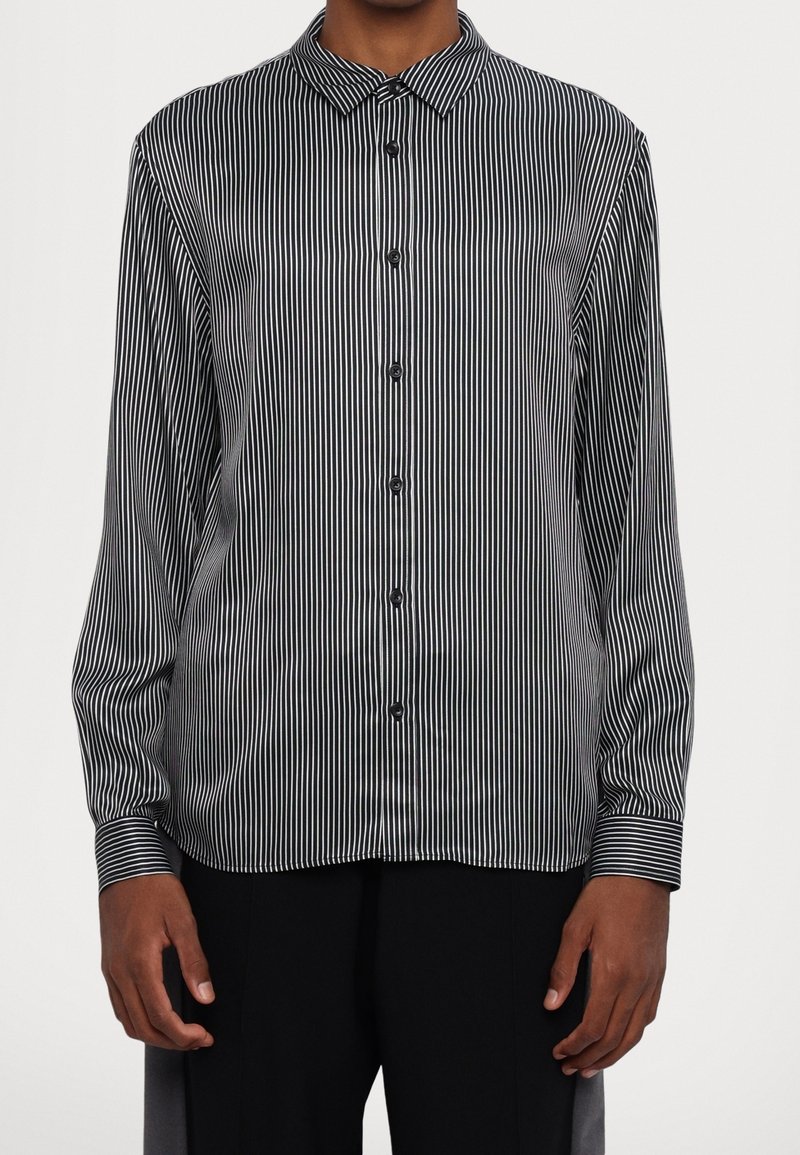 Homme portant une chemise à rayures verticales noires et blanches à manches longues avec col et pantalon noir.