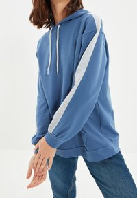 Persona vistiendo una sudadera azul con rayas de mangas gris claro y jeans azules, con las manos entrelazadas frente a un fondo blanco.
