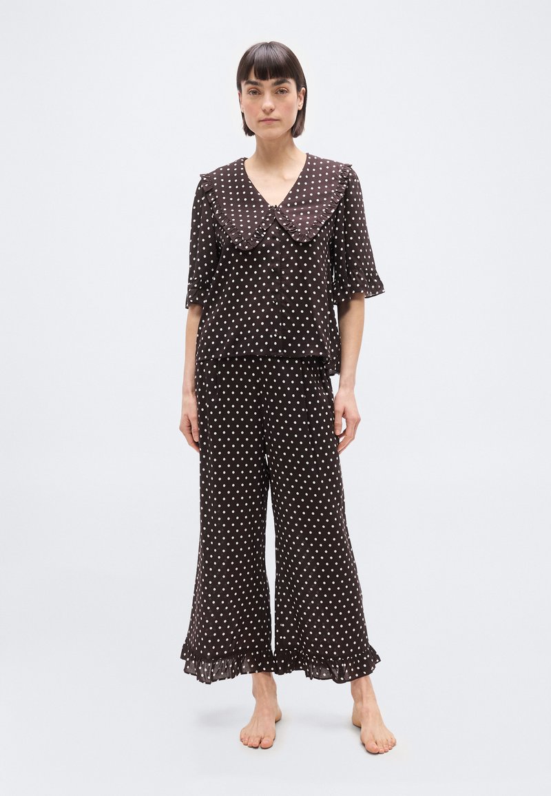 Femme portant un ensemble de pyjama noir à pois blancs avec un grand col à volants et un pantalon à jambes larges, debout pieds nus.