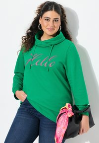 Felpa verde con collo a cowl e testo "Hello" in rosa, abbinata a jeans scuri e una borsa nera accentuata da una sciarpa rosa e arancione.