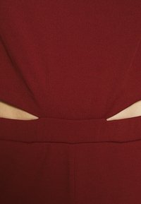 Gros plan sur un vêtement rouge foncé avec des découpes symétriques à la taille révélant la peau des deux côtés.