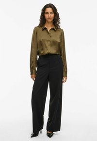 Camicia di satin verde oliva con colletto, maniche lunghe e pantaloni neri su misura, abbinati a décolleté nere lucide.