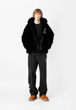 Chaqueta de piel sintética negra con un cuello ancho, combinada con pantalones negros. Detalles destacables: acentos bordados y mocasines naranjas.