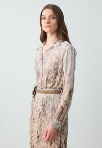 Sheer long-sleeve dress with a snake print pattern in beige and brown tones. Features a collared neckline and a belted waist with a wide buckle.