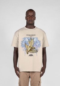 Beige bomull T-skjorte med grafisk design som viser en fugl, blå skyer, og teksten 'VINTAGE DREAMS' over og 'SEASON' under.