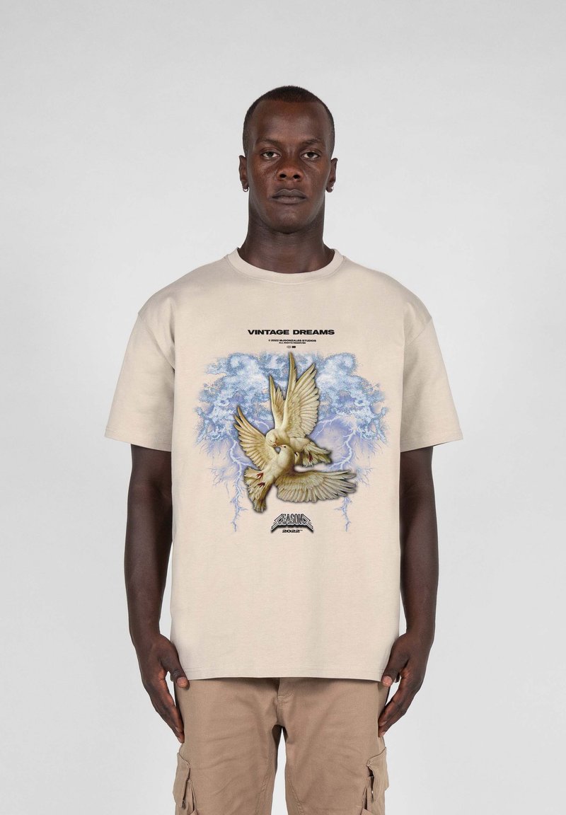 Beige bomull T-skjorte med grafisk design som viser en fugl, blå skyer, og teksten 'VINTAGE DREAMS' over og 'SEASON' under.