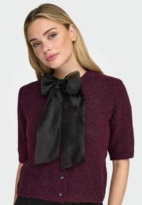 Cardigan bordeaux lavorato a maglia con maniche corte, caratterizzato da un grande fiocco nero in raso sul collo e chiusura con bottoni sul davanti.