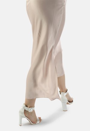 Eine Frau spaziert in einem hellrosa satin Midi-Rock und weißen, hochhackigen Sandalen mit Knöchelriemen vor einem einfarbigen Hintergrund.