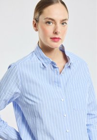 Chemise boutonnée bleu clair avec des rayures blanches verticales, col, manches longues, poche sur la poitrine gauche et fermeture à boutons sur le devant.