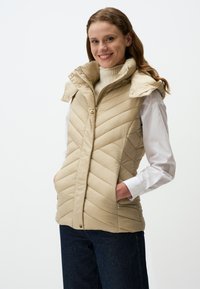 Beige gesteppte Weste mit hohem Kragen, Druckknöpfen und diagonaler Naht. Getragen über einem cremefarbenen Rollkragenpullover, kombiniert mit dunklen Jeans.