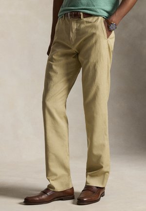 Muž oblečený v béžových chinos nohaviciach, hnedom koženom opasku, hnedých kožených mokasínach, zelenej košeli a náramkových hodinkách s koženým remienkom, stojaci s rukami v kapse.