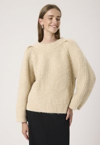 Sweter w kolorze kremowym, wykonany na drutach, z prążkowaną teksturą, charakteryzujący się luźnym krojem, szerokimi rękawami i wysokim dekoltem.