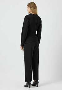 Haut noir à manches longues avec un col rond et un zip dans le dos, associé à un pantalon noir taille haute et des chaussures à talons noires.