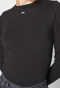 Maglietta a maniche lunghe nera in materiale elasticizzato, con un piccolo logo rosso, bianco e blu sul collo. Tessuto testurizzato.