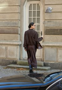 Femme en robe et veste marron foncé porte un sac à main et un café en marchant vers une porte claire installée dans un bâtiment en pierre.