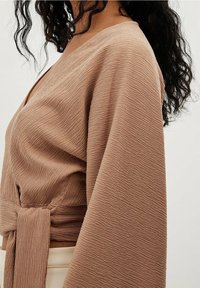 Brun tekstureret omslagstop med brede ærmer og en knyttet taljeparti. Har en dyb V-udskæring og ribbet stoftekstur.