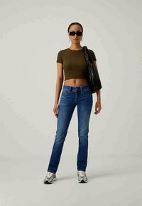 Jeans de denim azul de corte slim com desbotamento, emparelhados com um top cropped ajustado em verde oliva. Os acessórios incluem óculos de sol pretos e uma bolsa preta texturizada.
