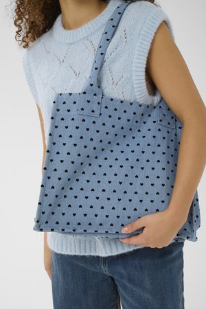 Donna che indossa un top senza maniche di maglia azzurro chiaro e jeans blu, che tiene una borsa a tracolla blu con motivi a cuore neri.