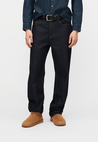 Σκούρα μπλε τζιν με αντίθετη ραφή, συνδυασμένα με ταμπά σουέτ μποτάκια αστραγάλου και μπλούζα από τζιν φορεμένη μέσα στο παντελόνι με μαύρη ζώνη.