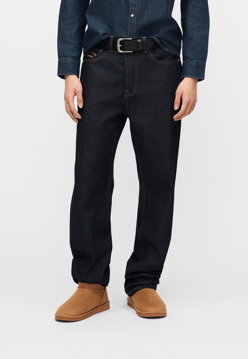 Σκούρα μπλε τζιν με αντίθετη ραφή, συνδυασμένα με ταμπά σουέτ μποτάκια αστραγάλου και μπλούζα από τζιν φορεμένη μέσα στο παντελόνι με μαύρη ζώνη.