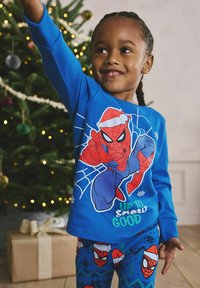 Bambino sorridente in pigiama delle festività di Spider-Man blu alza il braccio vicino a un albero di Natale decorato e a un regalo incartato.