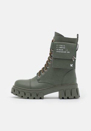 Bottes basses vertes à lacets avec une semelle épaisse et dentelée, des ornements décoratifs et une poche latérale avec des détails de texte imprimé.