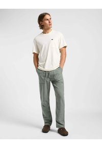 Vaaleanbeige lyhythihainen t-paita, jonka rinnassa on tasku, yhdistettynä vihreisiin joustovyötäröisiin housuihin ja ruskeisiin liukupolkimiin.