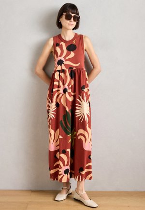 Maxi kjole i rust med store blomster mønstre i pink, grøn og sort; ærmeløs, A-formet design, parret med lyse flade sko.