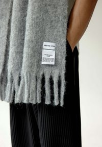 Écharpe en laine grise avec des franges, comportant une étiquette d'entretien suspendue. Écharpe présentée drapée sur un pantalon noir côtelé. Texture douce.