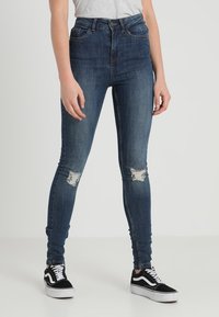 Jeans in denim a vita alta di colore blu scuro con toppe strappate sulle ginocchia. Abbinati a scarpe da ginnastica bianche e nere con un design classico.