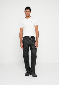 Bela majica s kratkimi rokavi z majhnim rdečim logotipom, kombinirana s črnimi usnjenimi hlačami z žepi in srebrno zaponko za pas. Model nosi črne superge.