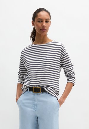 Femme aux cheveux bouclés attachés en queue de cheval, portant un t-shirt à manches longues rayé gris et blanc avec une poche à l'avant, et un pantalon bleu clair, les mains dans les poches.