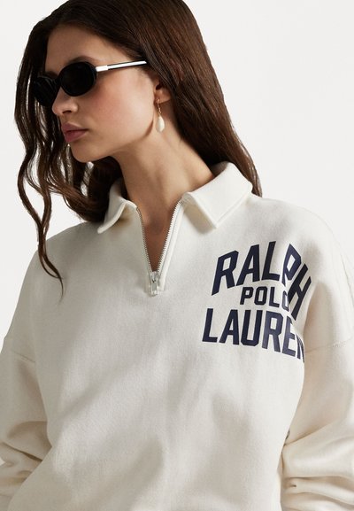 Valge lukuga sviiter, millel on "RALPH LAUREN" tumesiniste tähtedega. Kerge kangas, mugav lõige, varustatud krae ja soonikdetailidega. Mustad prillid.