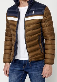 Homme portant une veste matelassée marron et bleu marine avec une bande blanche, partiellement zippée, par-dessus un t-shirt blanc et un jean bleu.