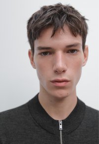 Veste bomber noire avec fermeture éclair et col rond, portée par une personne aux cheveux courts et en désordre sur un fond uni.