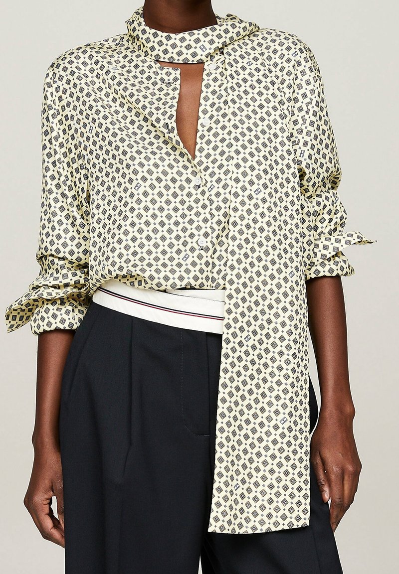 Blouse crème à manches longues avec motif losange noir, col montant avec détail foulard, associée à un pantalon noir taille haute et une ceinture blanche.