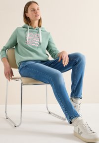 Sweat à capuche vert clair avec un imprimé graphique, associé à un jean bleu et des baskets blanches. Le mannequin est assis sur une chaise minimaliste en chrome.