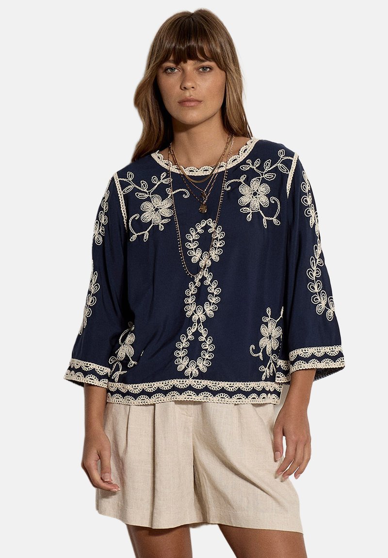 Femme portant un chemisier bleu marine avec une broderie florale blanche et un short plissé beige, accessoirisée avec des colliers superposés.