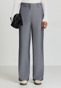 Persona che indossa pantaloni grigi larghi, una maglietta bianca a maniche lunghe con coste, sneakers bianche, mentre tiene una pochette di pelle nera con la mano sinistra.