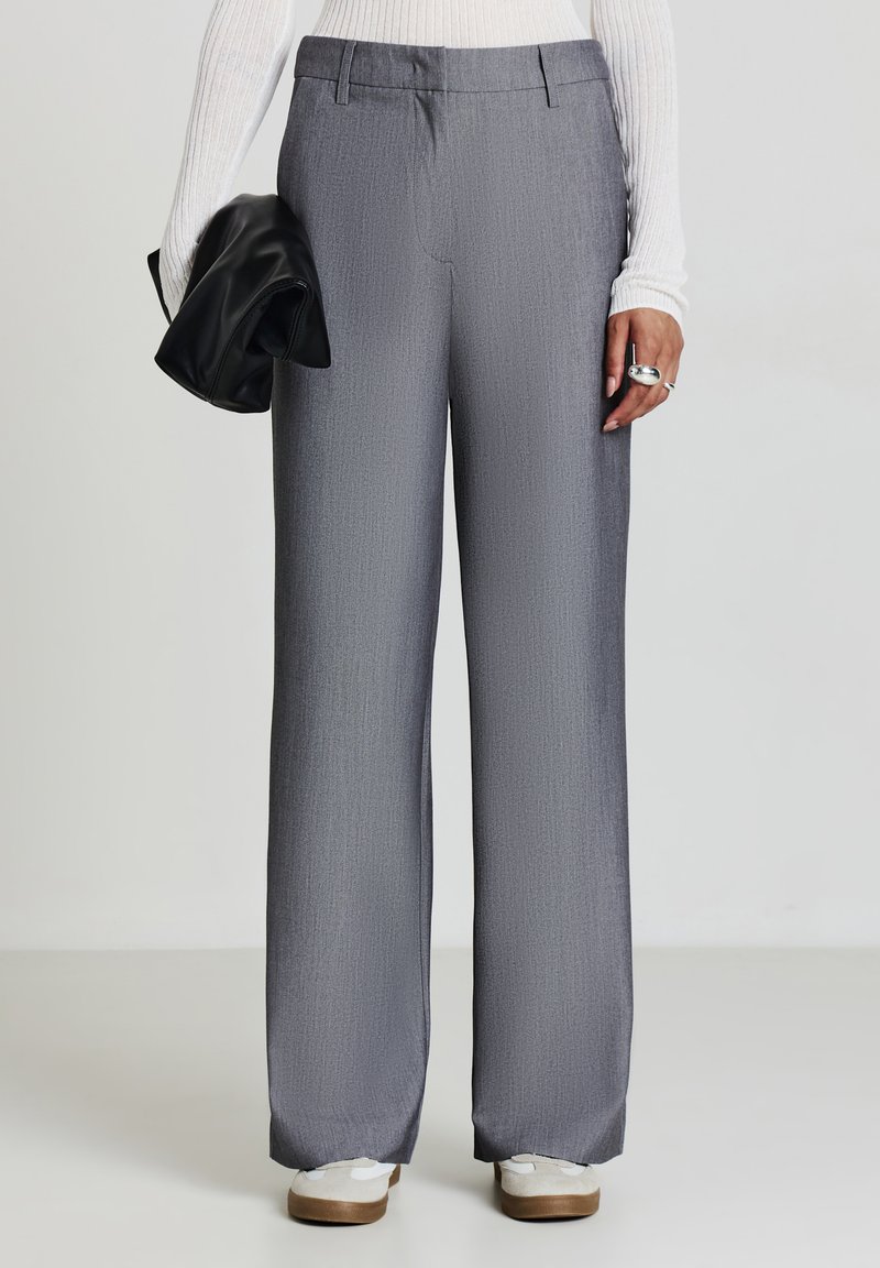 Persona che indossa pantaloni grigi larghi, una maglietta bianca a maniche lunghe con coste, sneakers bianche, mentre tiene una pochette di pelle nera con la mano sinistra.