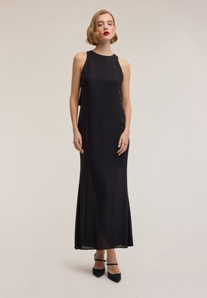 Abito da sera nero con scollo all'americana, lungo fino a terra, in tessuto leggero, vestibilità aderente con orlo svasato, abbinato a scarpe nere con punta appuntita.