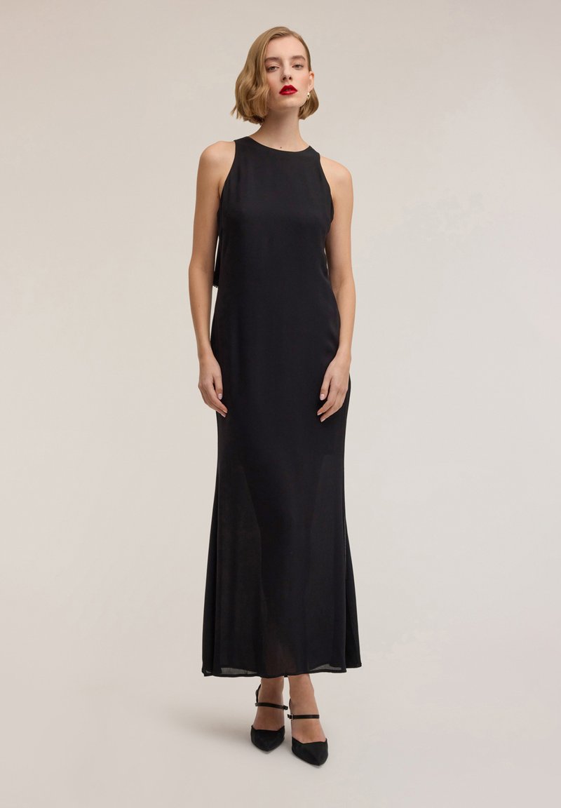 Abito da sera nero con scollo all'americana, lungo fino a terra, in tessuto leggero, vestibilità aderente con orlo svasato, abbinato a scarpe nere con punta appuntita.