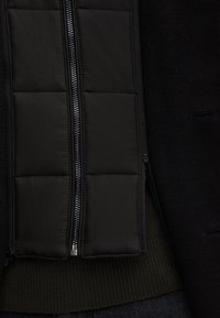 Zwarte jas met een doorgestikte, gevoerde binnenkant, grijze rits en elastische achterkant. Glad buitenmateriaal met een modern ontwerp.