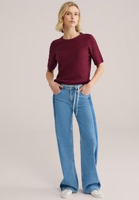 Kobieta w bordowym teksturowanym topie z krótkim rękawem oraz wysokim stanem szerokich niebieskich dżinsów z jasnym paskiem denimowym, stojąca z rękami w kieszeniach.