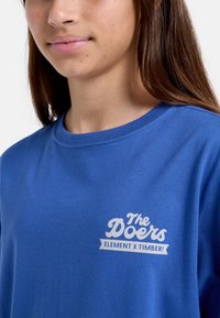 Camiseta de algodón azul con cuello redondo y un estampado gráfico blanco que dice "The Doers ELEMENT X TIMBER!" en el lado izquierdo.