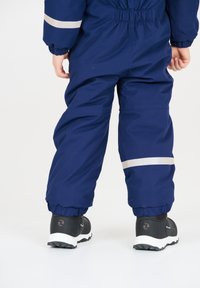 Marineblauw kinderwinteroverall met elastische manchetten, reflecterende zilveren strepen en een stevige rits. Zwarte sneakers met witte zolen.