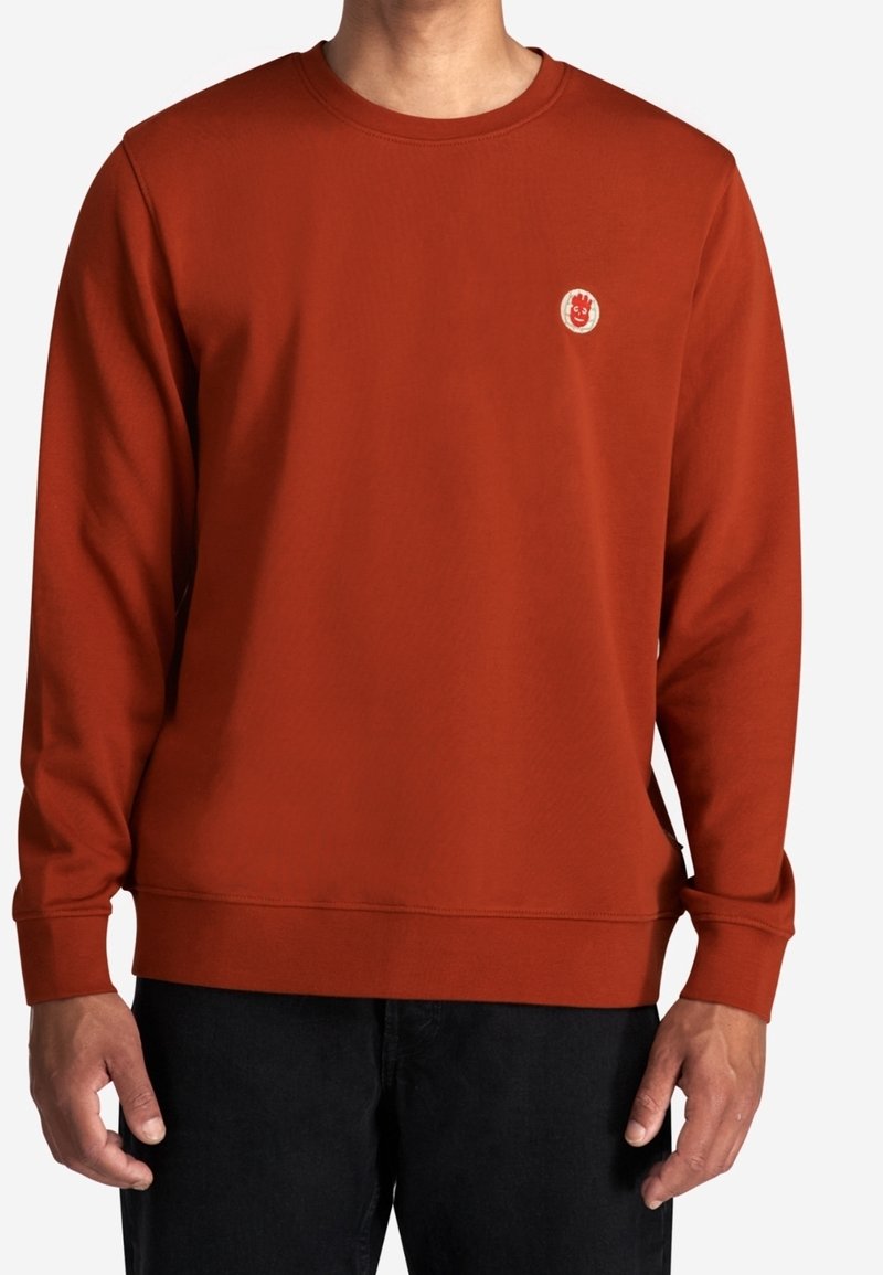 Homme portant un sweat-shirt à col rond manches longues de couleur rouille avec un petit logo rond sur le côté gauche de la poitrine et un pantalon noir.